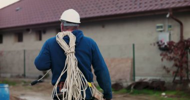 İnşaat alanında omuz üzerinde paketlenmiş elektrik kabloları taşırken, miğferli işçi araçları yönetirken ve kurulum için hazırlanırken binaya doğru yürüyordu.