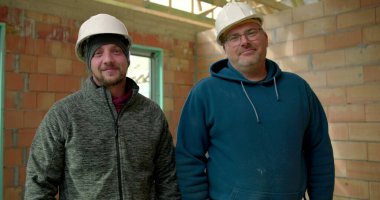 İnşaat halindeki bir binanın içinde gülümseyen iki inşaat işçisi. Birinin kolları bağlı, kısmen inşa edilmiş bir yapıda takım çalışması ve kendine güveni var.