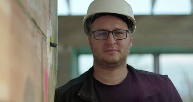 Başlıklı inşaat işçisi ciddi bir ifadeyle doğrudan kameraya bakıyor, inşaat alanında duruyor, duvara yaslanıyor.