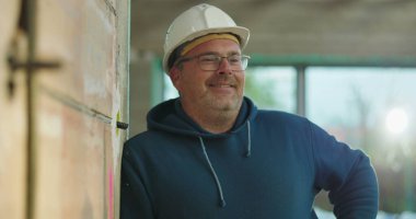 Sert şapkalı inşaat işçisi hafif bir gülümseme ile inşaat alanında duvara yaslanmış, rahat duruş, çevreyi gözlemleme, kapalı alan projeleri.