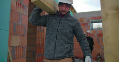 Omuzlarında ahşap kalaslar taşıyan iki inşaat işçisi, konut inşaatında takım çalışması, odaklanmış ifadeler, tuğla duvarlar ve iskelet yapısı