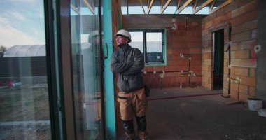 Genç inşaat işçisi, cam baret takılı, içinde inşaat malzemeleri ve açık tavan kirişleriyle dışarıda gözetleme yapıyor.