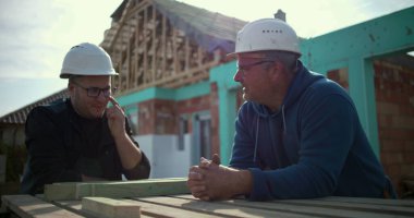 İnşaat alanında sohbet eden iki inşaat işçisi proje detayları, konut inşaatında takım çalışması ve işbirliği, arka planda ev iskeleti tartışmaları yapıyorlar.