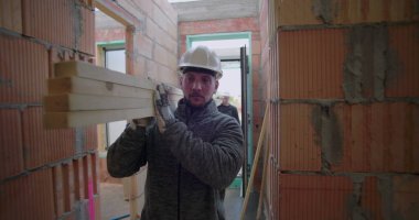 İnşaat işçisi, tamamlanmamış iskan binasının koridorundaki ahşap kalasları kaldırıyor. İşgücü ve inşaat alanında hassasiyet odaklanıyor.