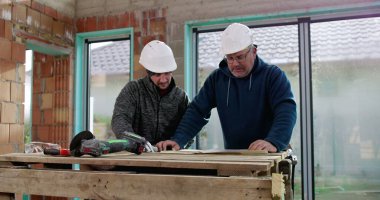 Sert şapkalı iki inşaat işçisi geçici ahşap bir masanın planlarını inceliyor, takım çalışması, planlama, bina inşaatı, işbirliği.