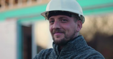 Kasketli inşaat işçisi kendine güvenerek gülümsüyor, şantiyede duruyor, arka planda açık çerçeve yapısını kuruyor, işçilikten tatmin ve gurur duyuyor