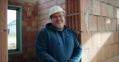 İnşaat işçisi tamamlanmamış binanın içinde duruyor, tuğla duvara yaslanıyor, güvenlik kaskı ve gözlük takıyor, kollar çapraz, kendinden emin ve cana yakın.