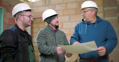 İnşaatçılar, bina planlarını birlikte gözden geçiriyorlar. İnşaat planlama, miğferli üç adam tamamlanmamış yapı projesini tartışıyorlar.
