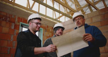 İnşaat işçileri kısmen inşa edilmiş yapının planlarını inceliyor, geniş bir inşaat alanında fikir alışverişinde bulunuyor ve proje detaylarını koordine ediyorlar.