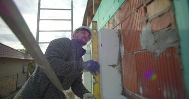 İzolasyon paneli ruh seviyesiyle karşılaştırılıyor. İnşaat işçisi yapı iskelesinde güvenlik ekipmanlarıyla çalışıyor. Duvarın dış duvarına tam oturmasını sağlıyor.