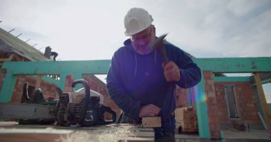 Ahşap bloğa çivi çakan inşaatçı, göreve odaklanmış, derme çatma tezgahın yanındaki testere, arka planda tamamlanmamış bina yapısı olan inşaat alanı.