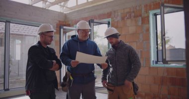 Üç inşaat işçisi planlarını gözden geçiriyorlar, güvenlik kaskı takıyorlar, proje detaylarını tartışıyorlar, takım çalışması ve bitmemiş endüstriyel alanda işbirliği yapıyorlar.