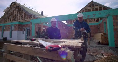 Birlikte çalışan iki inşaatçı, tezgahta testere, ahşap talaşı ve aletleri olan inşaat alanı, arka planda tamamlanmamış ev yapısı, projeye odaklanmış.