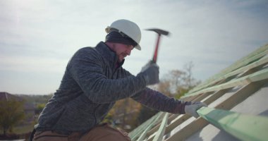 İnşaat işçisi çatıya tahta kirişler çakıyor, kask ve eldiven takıyor, çatı iskeletindeki işçilik ve işçiliğe odaklanıyor, konut projesine odaklanıyor.