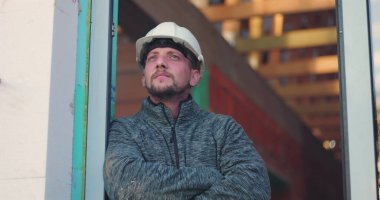 Kapıda inşaat işçisi, kollar çapraz, kasketli yana bakıyor, bina arka planı tamamlanmamış, kendini adamışlık, profesyonellik ve özen gösterme