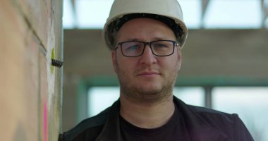 Başlıklı inşaat işçisi ciddi bir ifadeyle doğrudan kameraya bakıyor, inşaat alanında duruyor, duvara yaslanıyor.