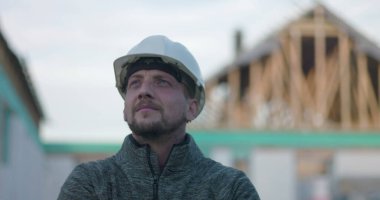 Başlıklı inşaat işçisi, düşünceli bir şekilde şantiyeye bakıyor, bina yapısının geçmişi, odak, planlama ve endüstriyel ithafı temsil ediyor.