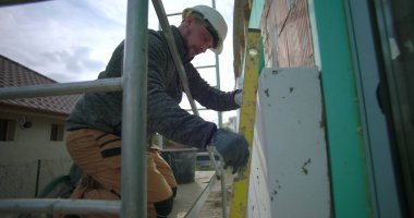 İnşaat işçisi duvardaki yalıtım panelini hizalamak için ruh seviyesini kullanıyor. Hassasiyet, güvenlik kaskı, arka planda yapı iskelesi ve açık hava projesine odaklanmış.