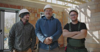 Üç inşaat işçisi kapalı alanda birlikte duruyorlar, güvenlik kaskı takıyorlar, bitmemiş bir inşaat sahasında gülümsüyorlar ve birlik gösteriyorlar, takım çalışmasını ve arkadaşlığı temsil ediyorlar.