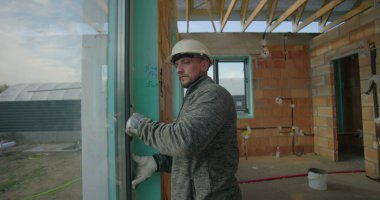 İnşaat işçisi inşaat teftişi sırasında pencere hizasını ve durumunu kontrol ediyor. Bitmemiş inşaat sahasında şapka ve güvenlik eldiveni giyiyor.