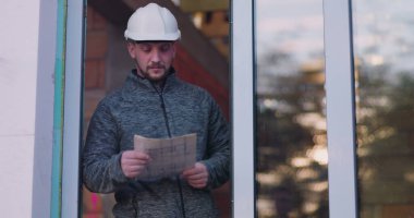 İnşaat işçisi kapının önünde planları tutuyor, planları inceliyor, başlık takıyor, tuğla duvar takıyor ve arka planda tamamlanmamış bina, odak ve inşaat planlamasını temsil ediyor.