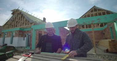 Tecrübeli müteahhitler bir inşaat sahasında ahşabı kalem ve mezurayla ölçüp işaretliyor, takım çalışması, hassasiyete, güvenlik kasklarına odaklanıyor, arka plandaki inşaat binasının altında.