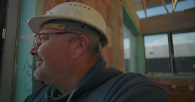 İnşaat işçisinin yakın çekimde dışarıdan bakan, tuğla duvar ve cam pencereli bitmemiş binanın içinde duran...