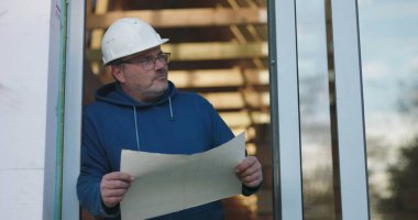 İnşaat işçisi inşaat planlarını tutuyor, şapka ve gözlük takıyor, planları incelemeye odaklanıyor, kapı eşiğinde duruyor, inşaat projesini denetliyor.