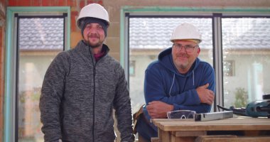 İnşaat işçileri inşaat halindeki çalışma tezgahının yanında gülümsüyor, miğferler takılı, takım çalışması ve inşaat sahasında dostluk, pozitif işyeri atmosferi