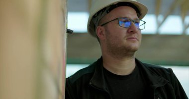 İnşaat işçisi kasketli, düşünceli bir şekilde yan tarafa bakıyor, inşaat alanında kapalı duruyor, duvara yaslanıyor, projeyi düşünüyor.