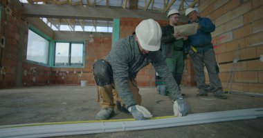 İnşaat işçisi, yerde mezura ile boruları ölçüyor, arka plandaki planları gözden geçiren iki meslektaşı, iç mimarideki takım çalışması ve planlama.