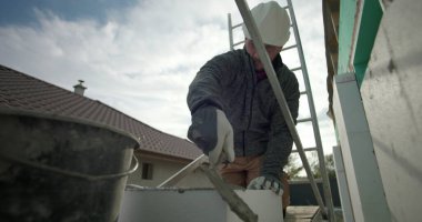 Havan topunu malayla hazırlamak, miğferli inşaat işçisi ve yapı iskelesinde eldiven, konut yenileme, duvarcılık malzemelerinin yakın çekim kullanımı
