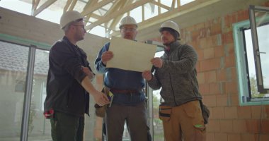 Miğferli inşaat işçileri planları analiz ediyor ve tamamlanmamış bir binanın içinde inşaat planlarını tartışıyor, proje elemanlarını işaret ediyor ve koordine ediyor.