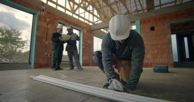 İnşaat işçisi, binanın iç kısımlarındaki PVC borularını diz çöküp ölçerken, arka plandaki meslektaşları da projeleri, inşaat alanındaki takım çalışmalarını gözden geçiriyorlar.