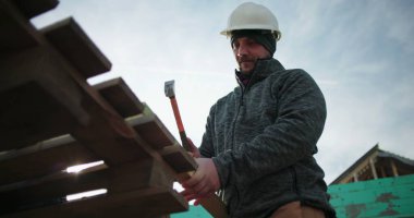 İnşaat işçisi çekiç kullanıyor, yakın çekim yapıyor, konut inşaatında marangozluk yapıyor, el aletine odaklanıyor, arka plandaki yapı iskeletine odaklanıyor.
