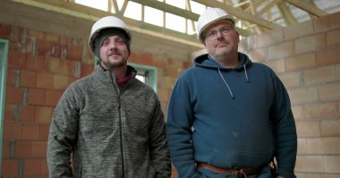 İnşaat alanında poz veren iki inşaat işçisi, güvenlik kaskı takan adamlar tamamlanmamış yapıda yan yana duruyorlar, takım arkadaşlığı ve iş gururu.