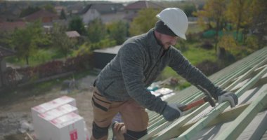 İnşaat işçisi çatıdaki tahta kirişleri çekiç, yan görüş, güvenlik kaskı ve eldiven ile sağlamlaştırıyor yerleşim alanındaki yüksek yapı üzerinde çalışıyor.