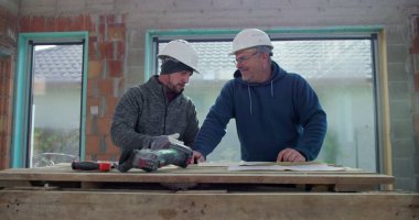 İki inşaat işçisi masa, takım çalışması ve planlama, kapalı inşaat sahası, başlık ve aletler, işbirliği çabası, inşa projesi üzerinde çalışıyor.