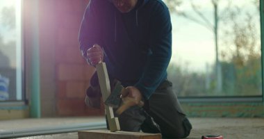 İnşaat işçisi, inşaat alanında diz çökerken ahşap çekiç kullanıyor. Etrafı tamamlanmamış tuğla duvarlarla çevrili malzemeleri korumaya odaklanıyor. İnşaat devam ediyor.