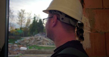 Dışarıya bakan inşaat işçisi, gülümseyen, yandan bakan, inşaat enkazı ve arka planda ağaçlar olan, gevşek ifadeli.