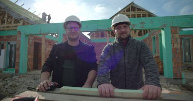 Yeni ev inşaatında iki erkek inşaatçı aletlerle ayakta duruyor, kameraya bakıyor, başlık takıyor, konut projesinde çalışıyor, çerçeveyi görünür kılıyor.