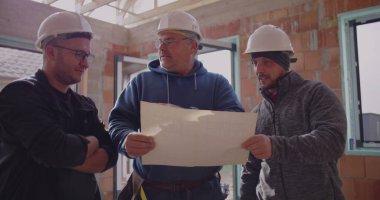 Üç inşaat işçisi planlarını gözden geçiriyorlar, güvenlik kaskı takıyorlar, proje detaylarını tartışıyorlar, takım çalışması ve bitmemiş endüstriyel alanda işbirliği yapıyorlar.