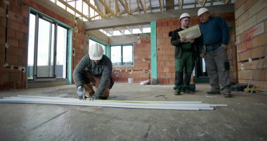 Kattaki PVC borularını ölçen inşaat işçisi, arka plandaki planları, iç inşaat düzenini, takım çalışmasını ve işbirliğini inceleyen iki meslektaşla birlikte