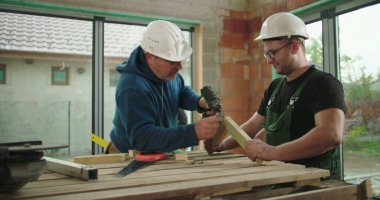 Tahta kiriş üzerinde elektrikli matkap kullanan iki inşaat işçisi, işbirlikçi ekip çalışması, inşaat projesi, güvenlik kaskları, inşaat alanında aktif çalışma ortamı