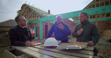 İnşaat ekibi pizza molasının keyfini çıkarıyor, üç inşaatçı şantiyede birlikte yemek yiyor, konut inşaatı sırasında öğle yemeği yiyor.