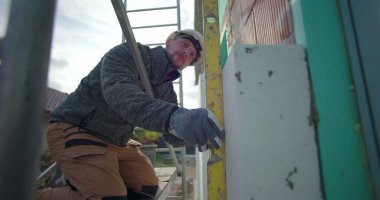 İzolasyon tesisatı üzerinde çalışan, iskele üzerinde diz çöken, kesinlik için seviye aletini kullanan, inşaat halindeki konut evinin dış duvarına odaklanmış bir yapı.