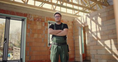 İnşaat işçisi güvenle duruyor. Kolları bağlı, kaskı ve tulumuyla inşaat alanında duruyor. İnşaat alanında gururlu ve profesyonel duruş sergiliyor.