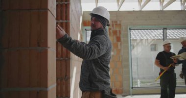 Duvarı kalemle ölçen inşaat işçisi, güvenlik miğferinde odaklanmış miğferle içeride tuğla yapı, hassasiyet ve detaylara dikkat ediyor.