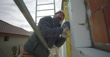 Yalıtım panelini ruh seviyesine ayarlıyorum. İnşaat işçisi iskan duvarına doğru yerleştirme, güvenlik teçhizatı görünür, tuğla zemin.