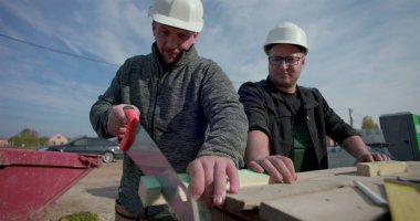 İki inşaatçı birlikte çalışıyor, biri odun kesmek için el testeresi kullanırken diğer saatler, etkin inşaat sahasında takım çalışması, yapı iskeleti ve aletler etrafında.